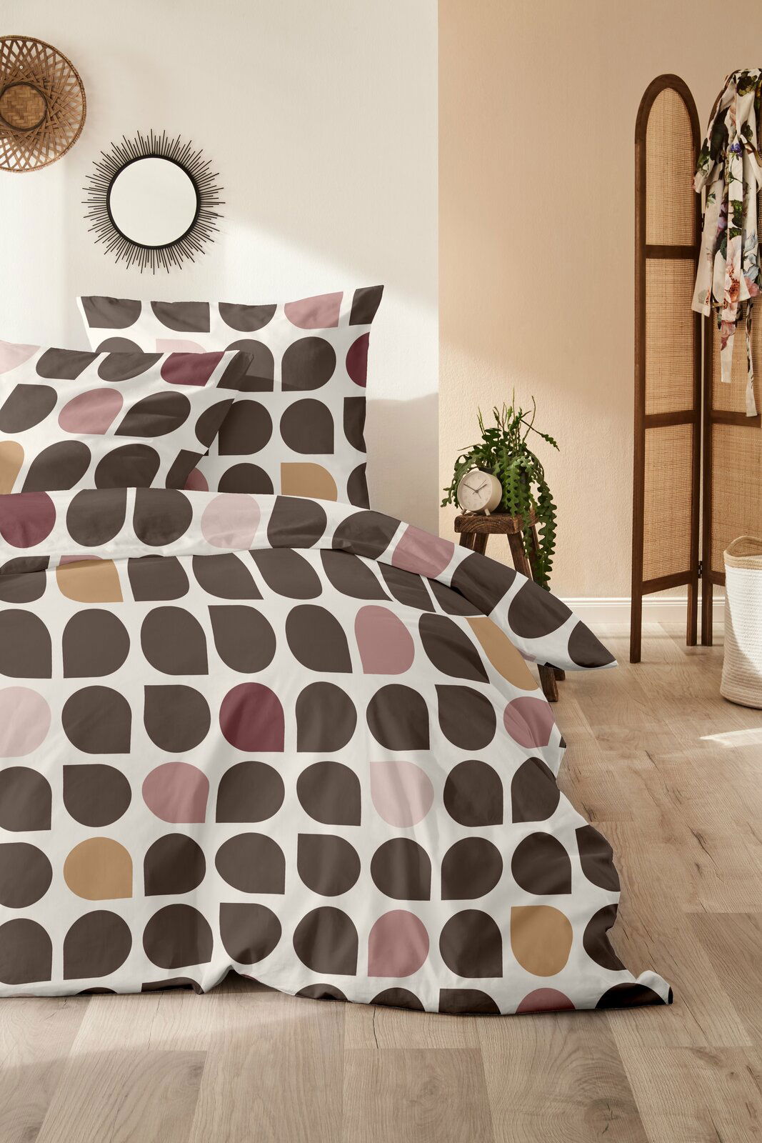 Bettwäsche mit geometrischem Muster in Braun-, Rosa- und Beigetönen, aus der Perspektive eines gemütlichen Schlafzimmers mit Holzboden und Dekoration.