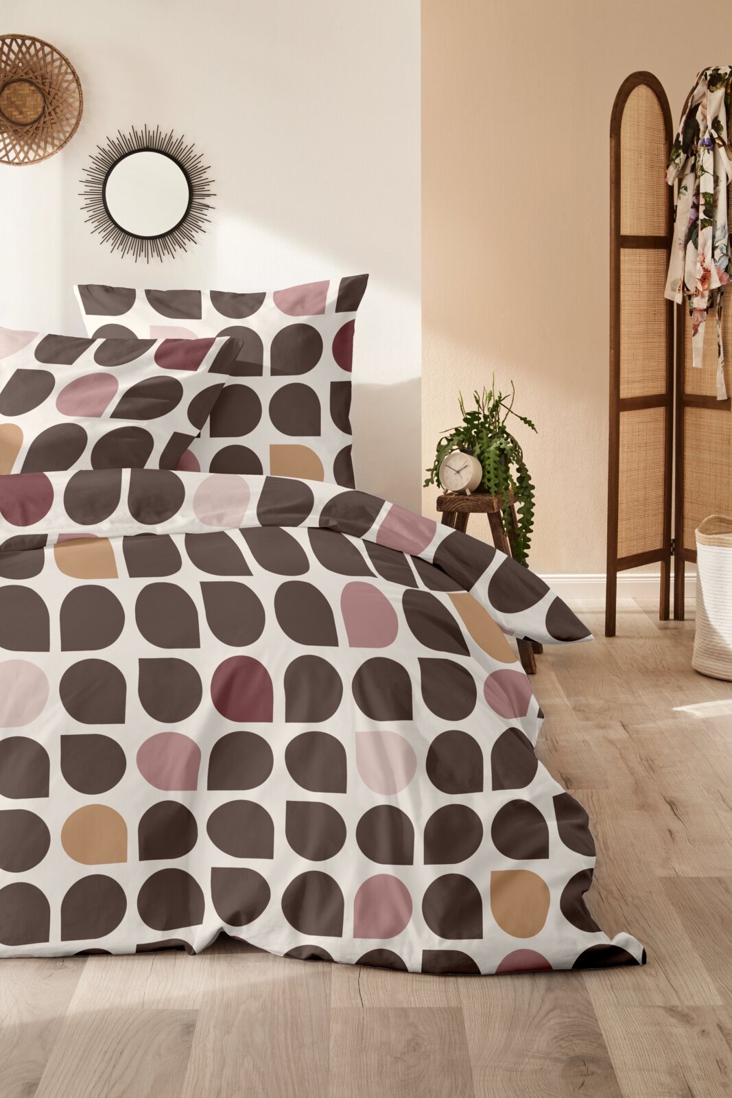 Bettwäsche mit geometrischem Muster in Braun-, Rosa- und Beigetönen, aus der Perspektive eines gemütlichen Schlafzimmers mit Holzboden und Dekoration.