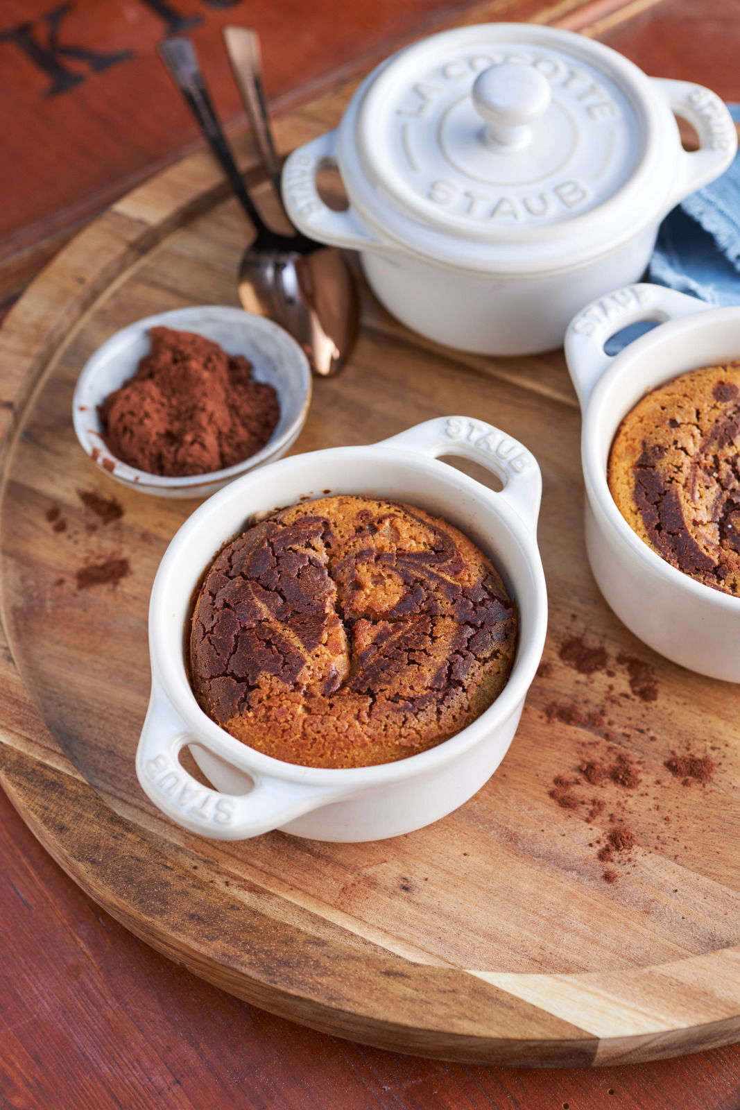 Vogelperspektive auf ein Set von elfenbeinfarbenen Mini-Cocottes mit gebackenem Dessert, umgeben von Kakaopulver und Löffeln auf einem Holztablett.