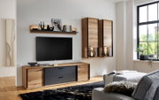 Moderne Wohnzimmer-Wohnwand in Eiche-Optik mit schwarzen Fronten, Lowboard, Wandregal und zwei Hängeschränken neben einem wandmontierten TV, fotografiert in frontaler leicht seitlicher Perspektive.