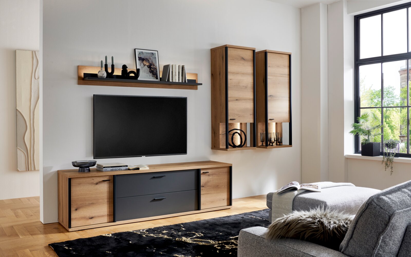 XORA Wandboard MELO Moderne Wohnzimmer-Wohnwand in Eiche-Optik mit schwarzen Fronten, Lowboard, Wandregal und zwei Hängeschränken neben einem wandmontierten TV, fotografiert in frontaler leicht seitlicher Perspektive.