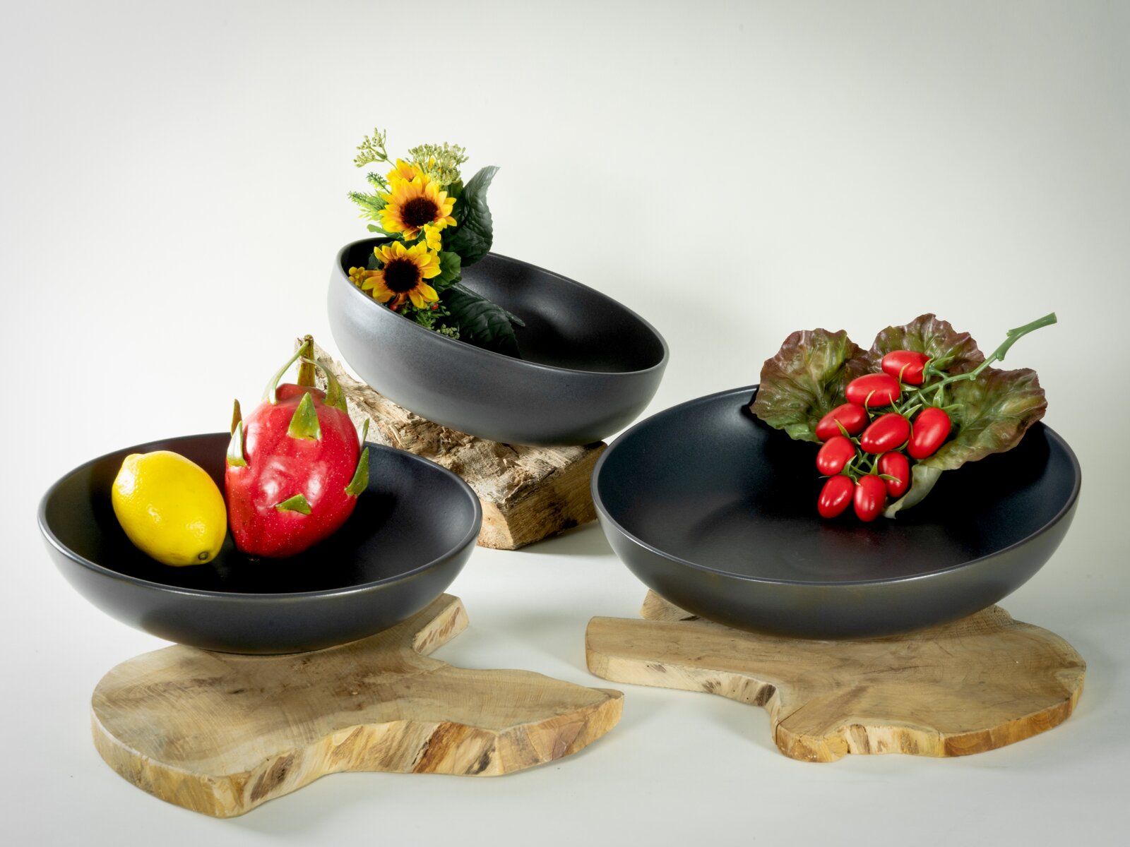 Drei matte schwarze Servierschalen in unterschiedlichen Größen auf Holzplatten, dekoriert mit Zitrone, Drachenfrucht, Tomaten und Blumen, in schräger Vorderansicht
