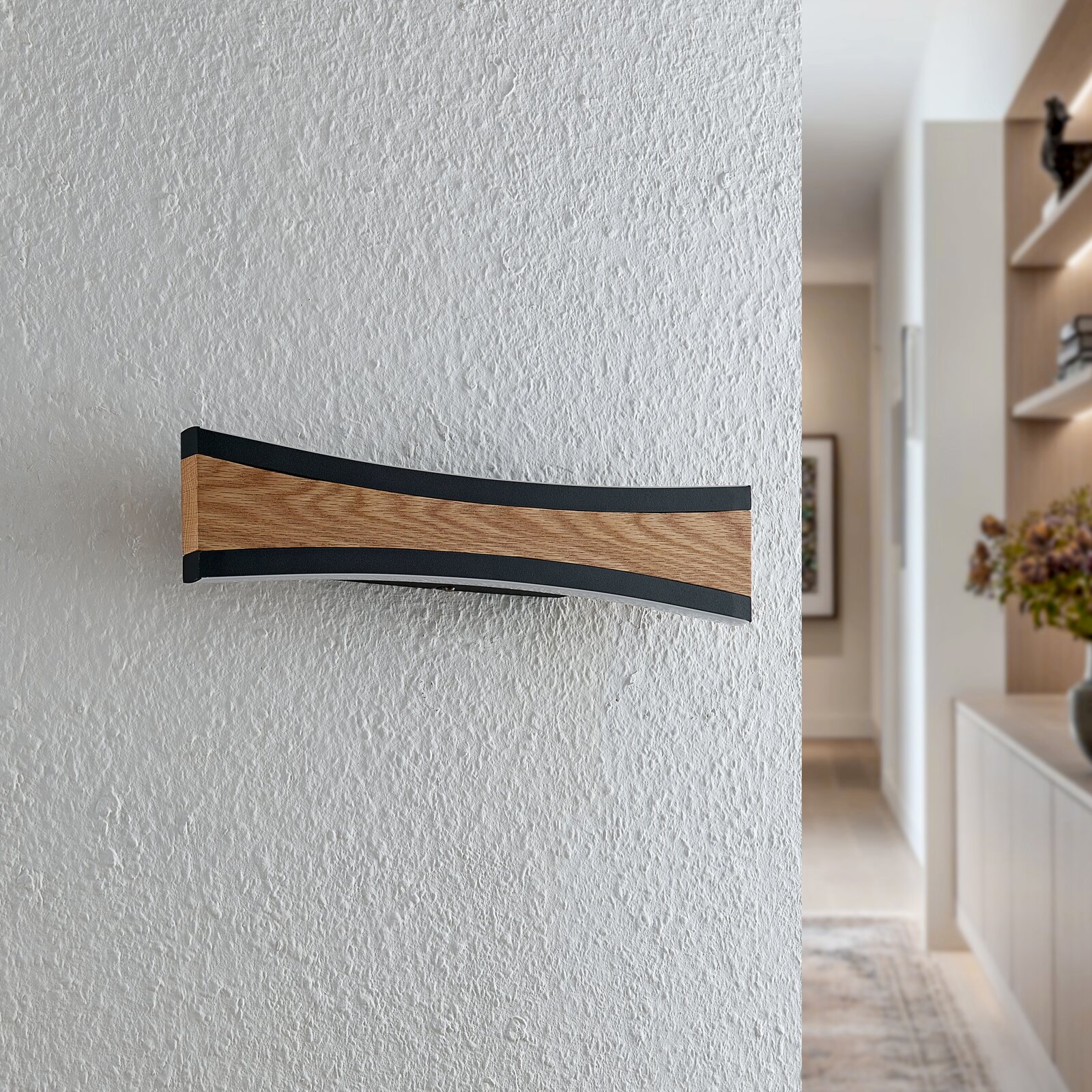 Moderne, geschwungene LED-Wandleuchte mit Holzoptik und schwarzem Rahmen an strukturierter weißer Wand im Flur, frontal aus leicht seitlicher Perspektive aufgenommen.