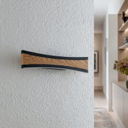 Moderne, geschwungene LED-Wandleuchte mit Holzoptik und schwarzem Rahmen an strukturierter weißer Wand im Flur, frontal aus leicht seitlicher Perspektive aufgenommen.
