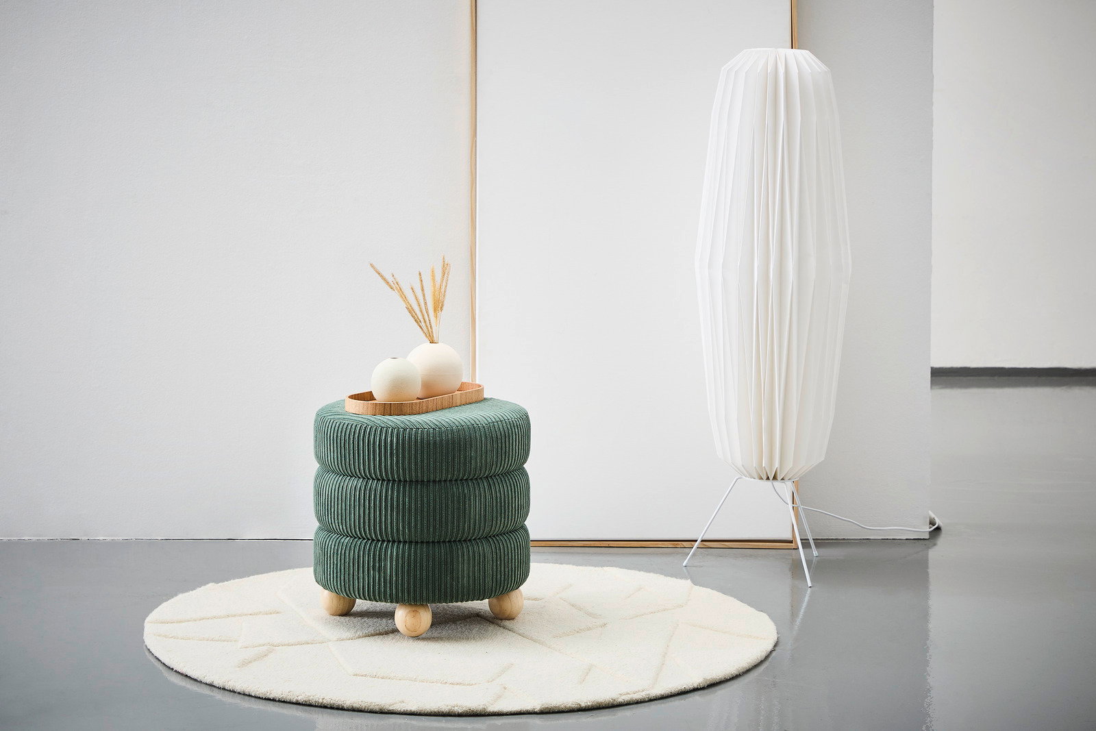 Grüner, runder Hocker mit Holzfüßen, dekoriert mit einem Tablett und Vasen, auf einem cremefarbenen Teppich, seitliche Perspektive.