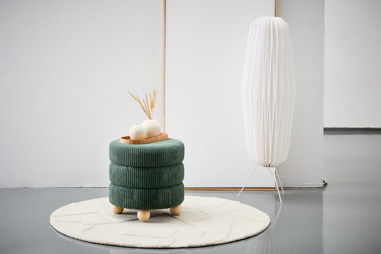XORA Hocker MOLLY Grüner, runder Hocker mit Holzfüßen, dekoriert mit einem Tablett und Vasen, auf einem cremefarbenen Teppich, seitliche Perspektive.
