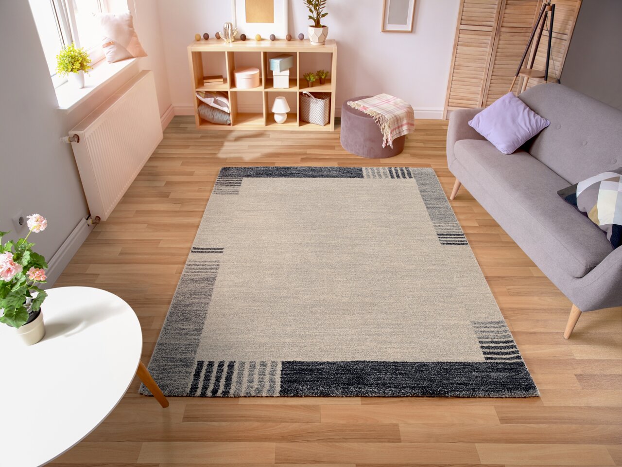Moderner Frisé-Teppich in einem stilvollen Wohnzimmer, von oben betrachtet. Der Teppich hat ein geometrisches Design in Grau- und Beigetönen und liegt auf einem hellen Holzboden. Umgeben von einem Sofa, einem kleinen Tisch und einem Regal.
