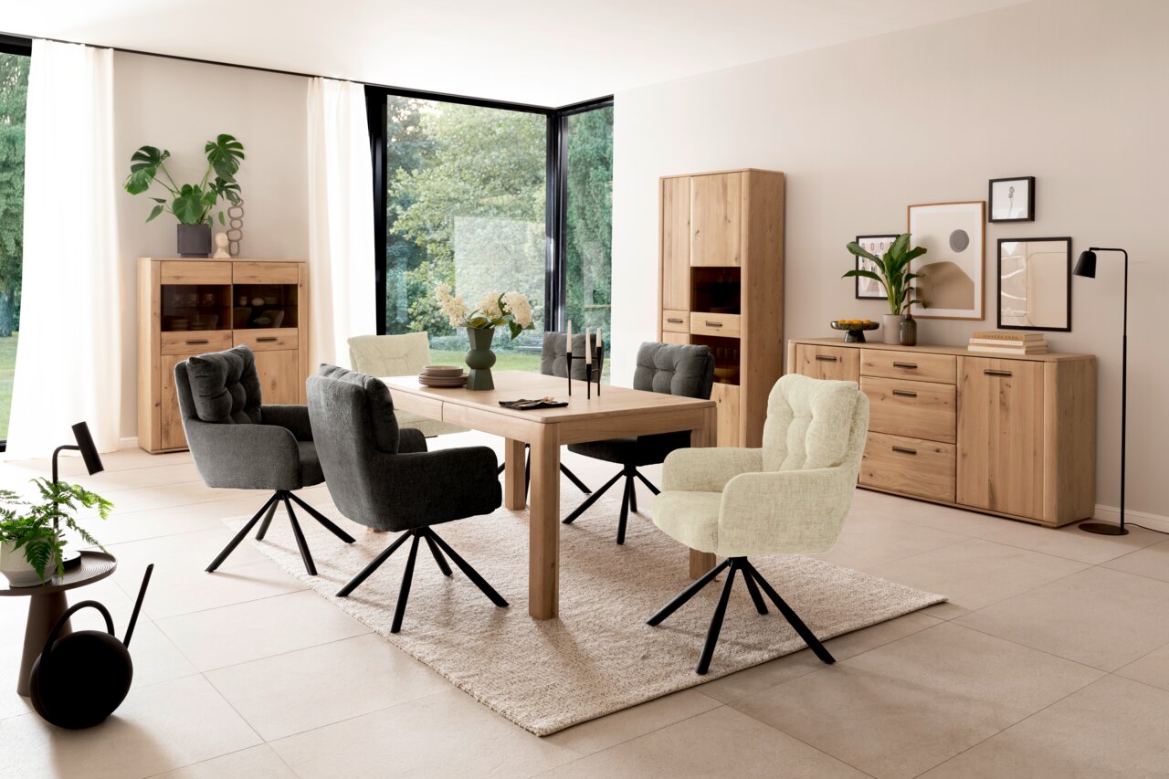 Esszimmer mit modernem Esstisch aus Holz, umgeben von gepolsterten Stühlen in Grau und Beige, mit Blick auf eine helle, von Pflanzen umgebene Fensterfront.