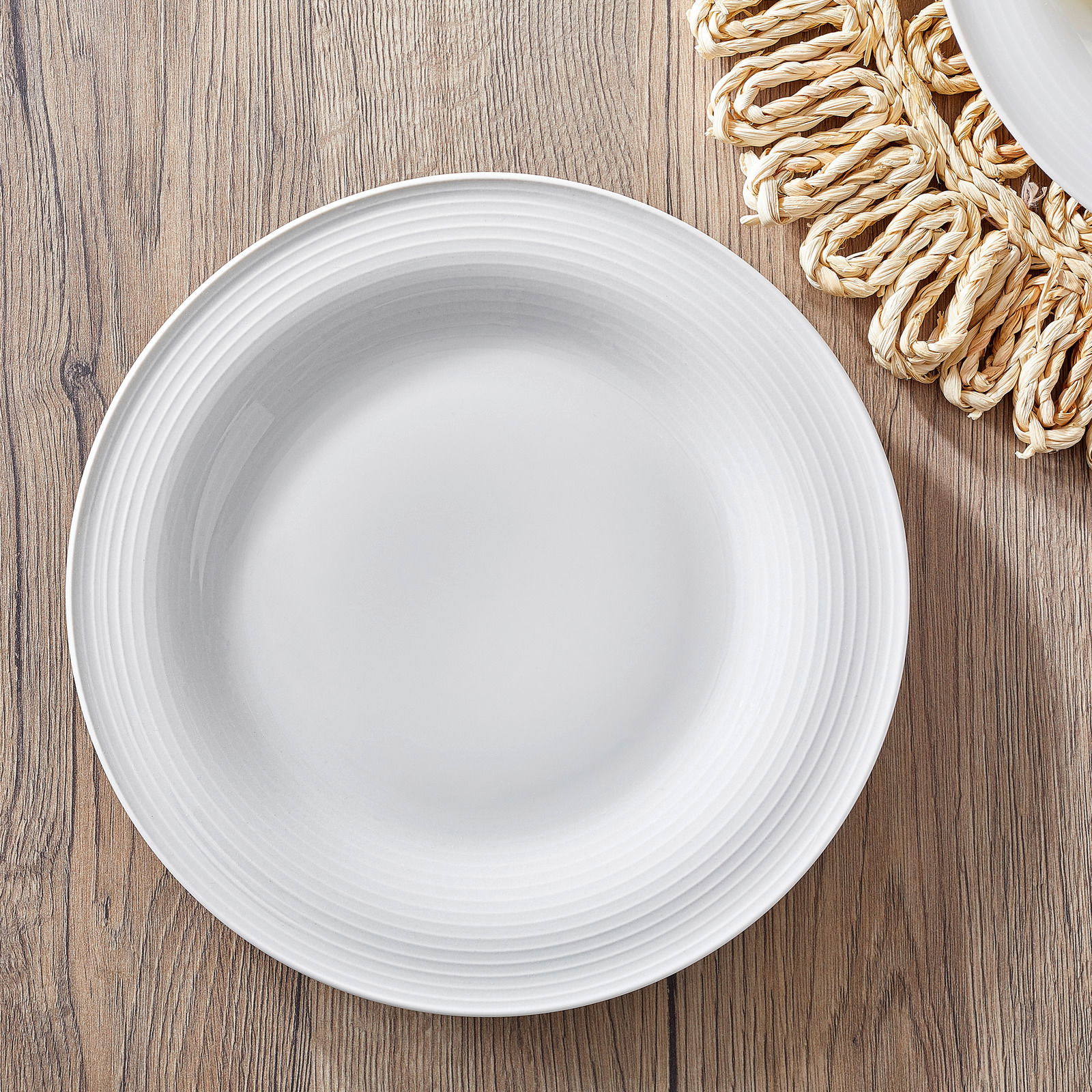 Weißer Teller mit feiner Rillenstruktur, von oben fotografiert, auf einem Holztisch mit geflochtenem Untersetzer.