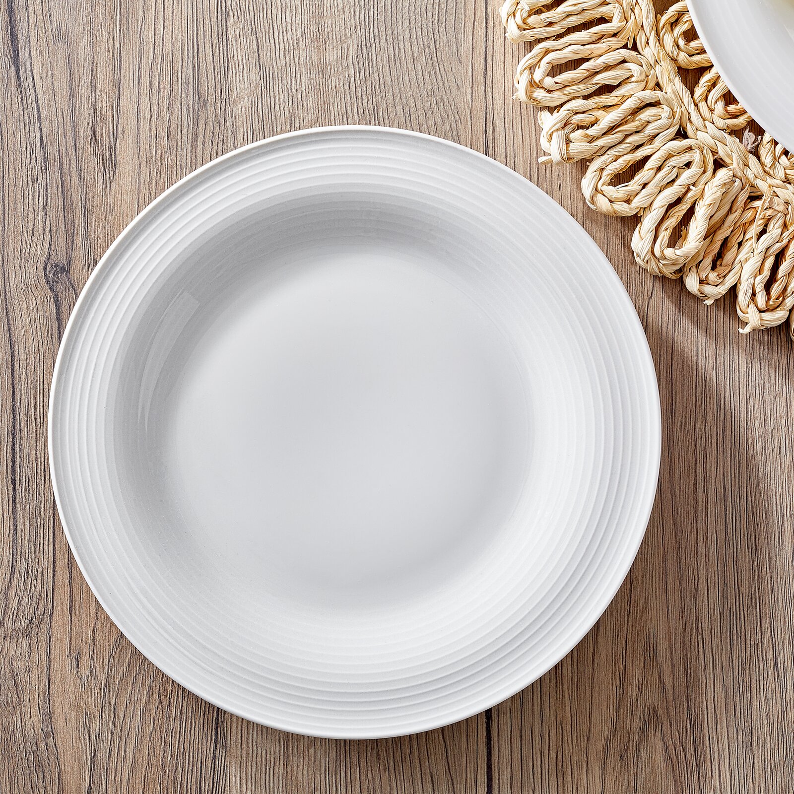Weißer Teller mit feiner Rillenstruktur, von oben fotografiert, auf einem Holztisch mit geflochtenem Untersetzer.