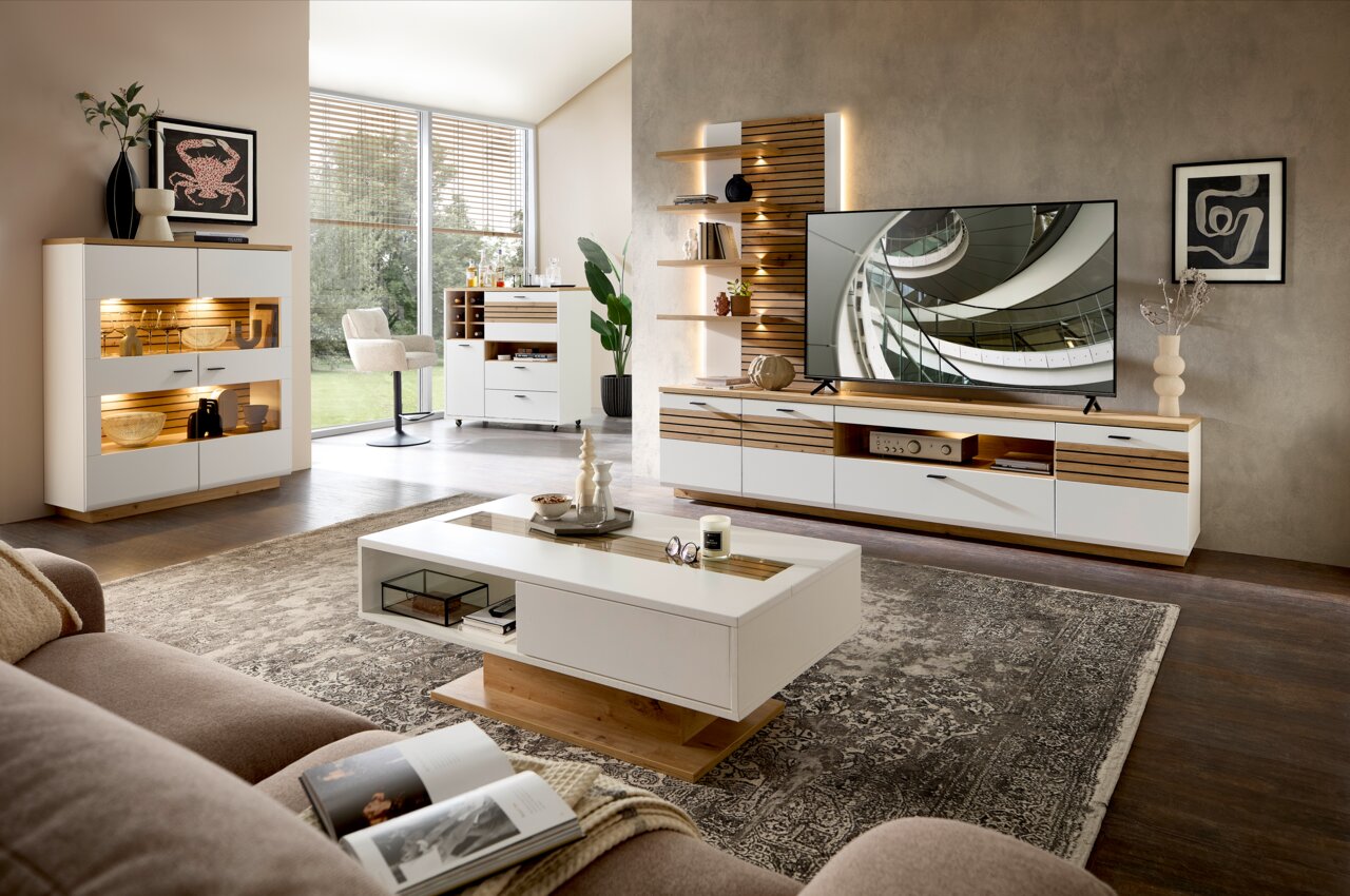 Moderne woonkamer met een stijlvol combielement als ruimteverdeler in wit en houtlook, gezien vanaf de voorkant. Geïntegreerde verlichting en decoratieve accessoires zorgen voor een gezellige sfeer.