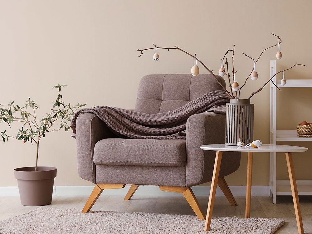 Gemütliche skandinavische Wohnzimmerdekoration zu Ostern mit Sessel, Osterzweigen im Kerzenhalter, Beistelltisch und Regal in Naturtönen