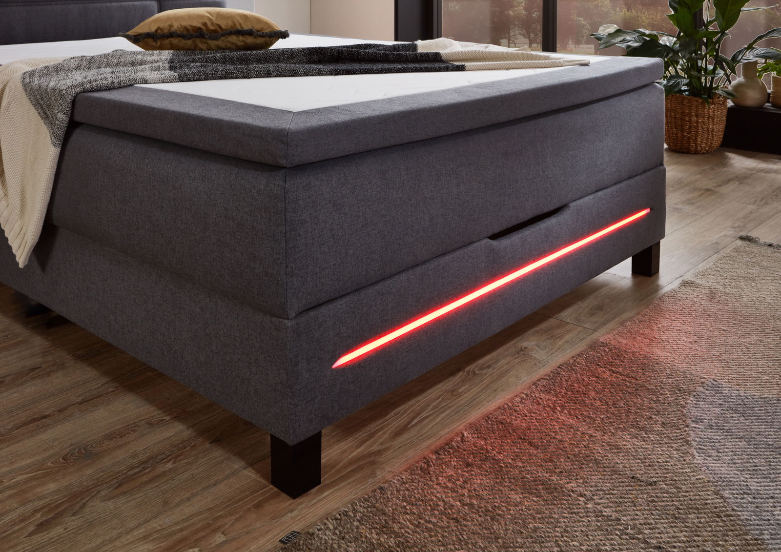Boxspringbett in grauem Stoff mit roter LED-Beleuchtung an der Seite, seitliche Perspektive