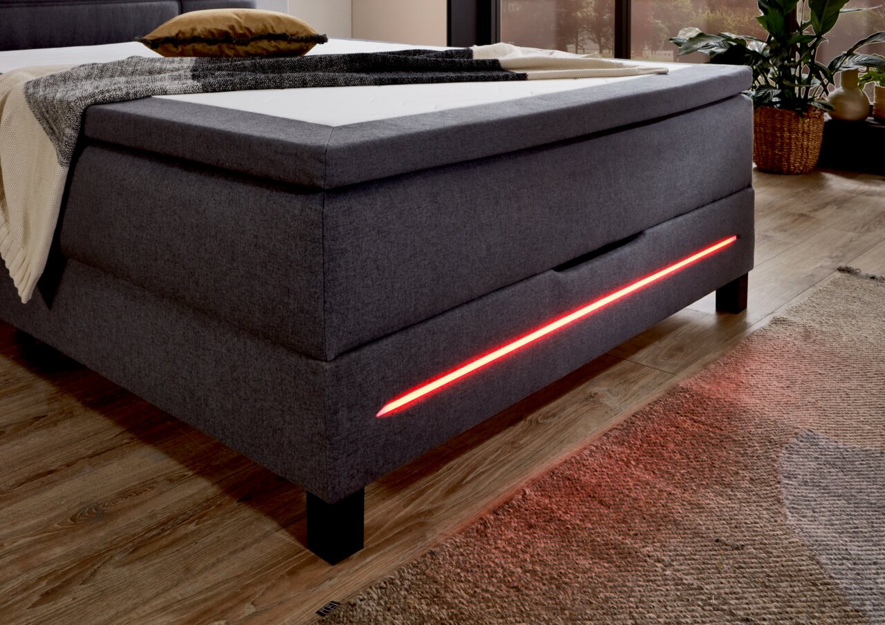 Boxspringbett in grauem Stoff mit roter LED-Beleuchtung an der Seite, seitliche Perspektive