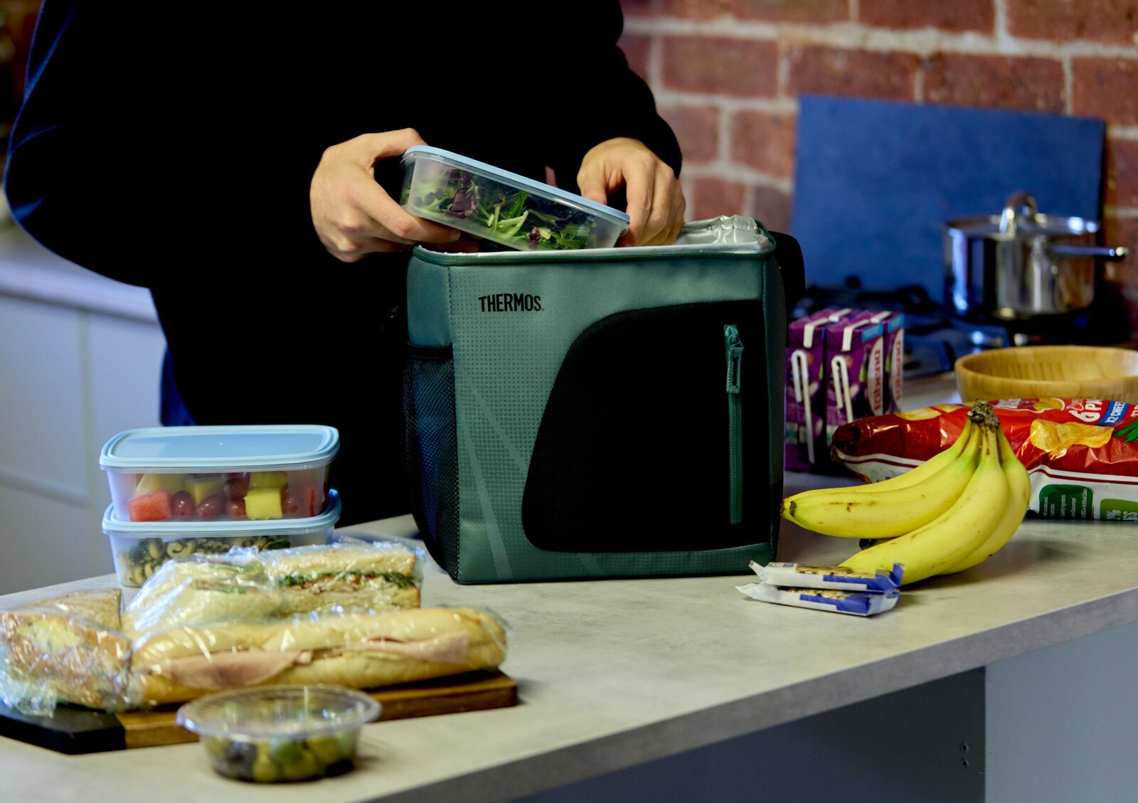 Thermos Kühltasche in Grün und Schwarz aus frontaler Perspektive, während sie in einer Küche mit einer Salatdose und weiteren Speisen befüllt wird