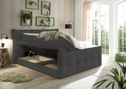 Boxspringbett mit Stauraum, seitliche Perspektive, modernes Design, grauer Stoffbezug, helle Raumgestaltung mit Pflanzen und großen Fenstern