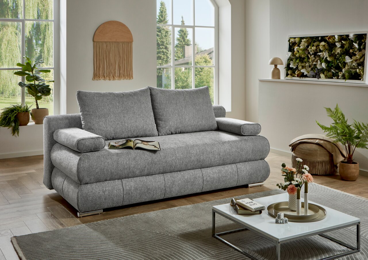 Graues Funktionssofa in einem modernen Wohnzimmer, frontal fotografiert, mit großen Fenstern im Hintergrund und dekorativen Pflanzen.