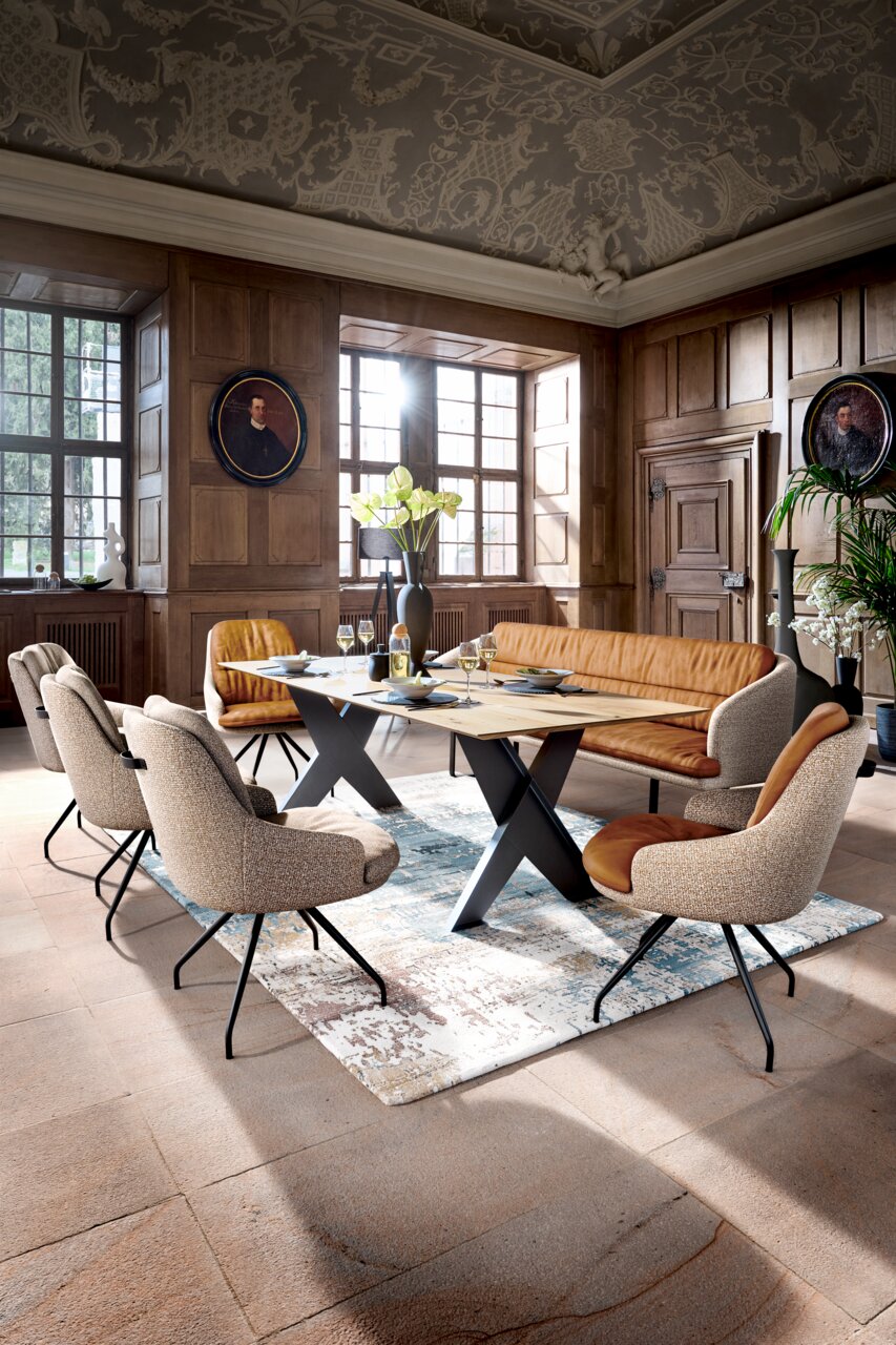 Dieter Knoll Fauteuil VERBRANDEN Moderne eetkamerset met een lichte houten tafel op een zwart X-vormig onderstel, gestoffeerde stoelen en cognackleurige zitmeubelen in een met hout beklede ruimte met grote ramen; perspectief: licht verhoogd vooraanzicht