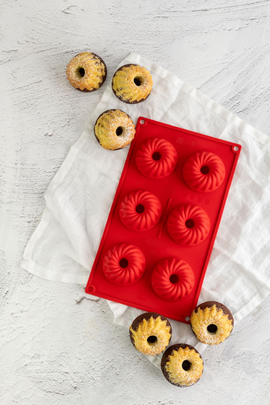Rote Silikon-Gugelhupfform für sechs Mini-Kuchen, von oben fotografiert, auf einem weißen Tuch liegend, umgeben von gebackenen Mini-Gugelhupfen.