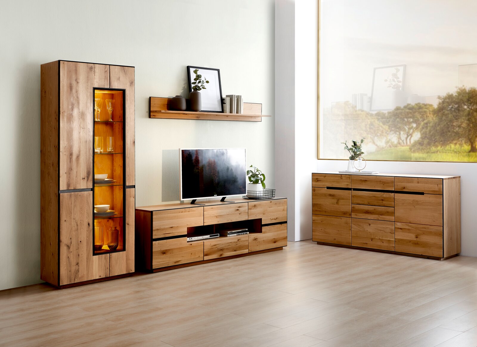Moderne woonkamermeubelset in massief houtlook met hoge vitrinekast met warm verlichte glazen deur, laag tv-meubel met lades en open vak, en brede dressoir; gefotografeerd in frontaal aanzicht met lichte hoekperspectief van links.
