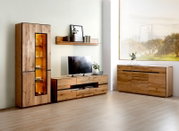 Moderne woonkamermeubelset in massief houtlook met hoge vitrinekast met warm verlichte glazen deur, laag tv-meubel met lades en open vak, en brede dressoir; gefotografeerd in frontaal aanzicht met lichte hoekperspectief van links.