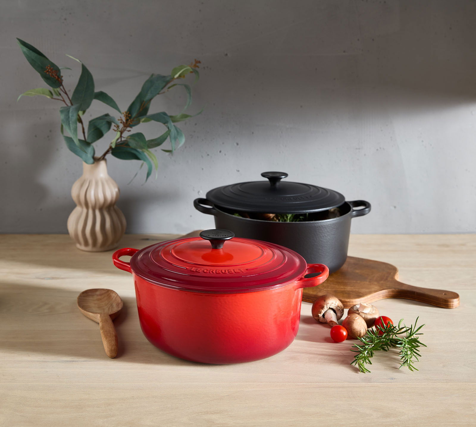 LE CREUSET Bräter 26cm Schwarz TRADITION Zwei runde Bräter in Kirschrot und Schwarz auf einem Holztisch, umgeben von frischen Kräutern und Gemüse, mit einer Vase im Hintergrund. Perspektive von vorne.