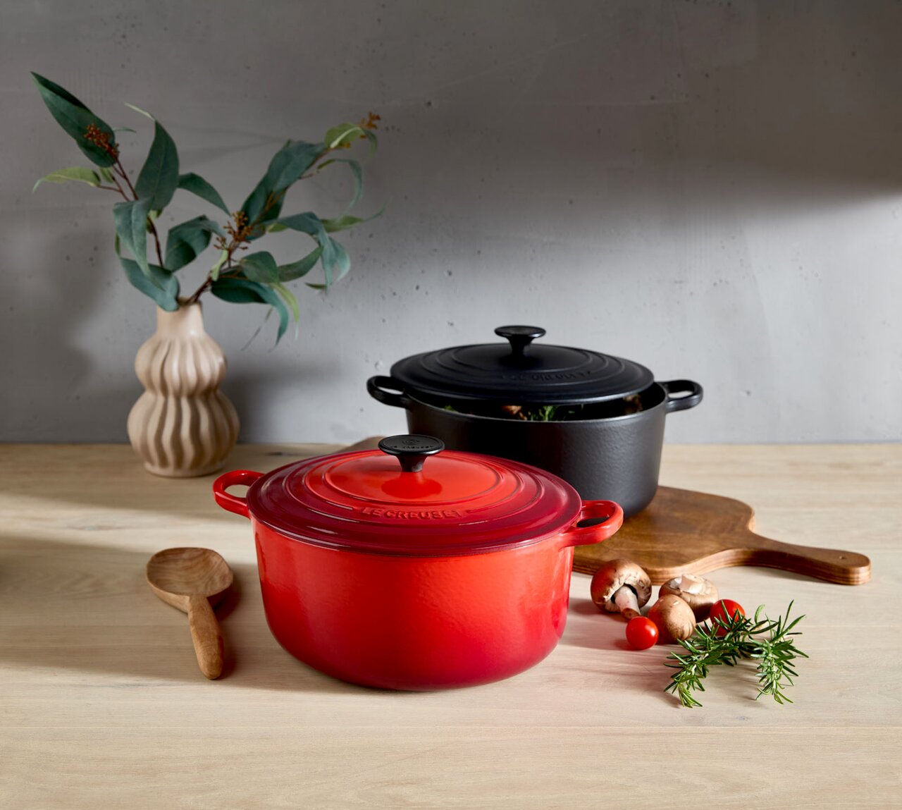 LE CREUSET Bräter 26cm Schwarz TRADITION Zwei runde Bräter in Kirschrot und Schwarz auf einem Holztisch, umgeben von frischen Kräutern und Gemüse, mit einer Vase im Hintergrund. Perspektive von vorne.