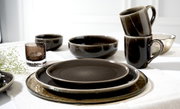 Verschiedene Geschirrteile aus der Ritzenhoff & Breker EDA UMBRA Serie, darunter Tassen, Schalen und Teller, in einer eleganten dunklen Farbgebung, auf einem Tisch arrangiert. Perspektive von schräg oben.