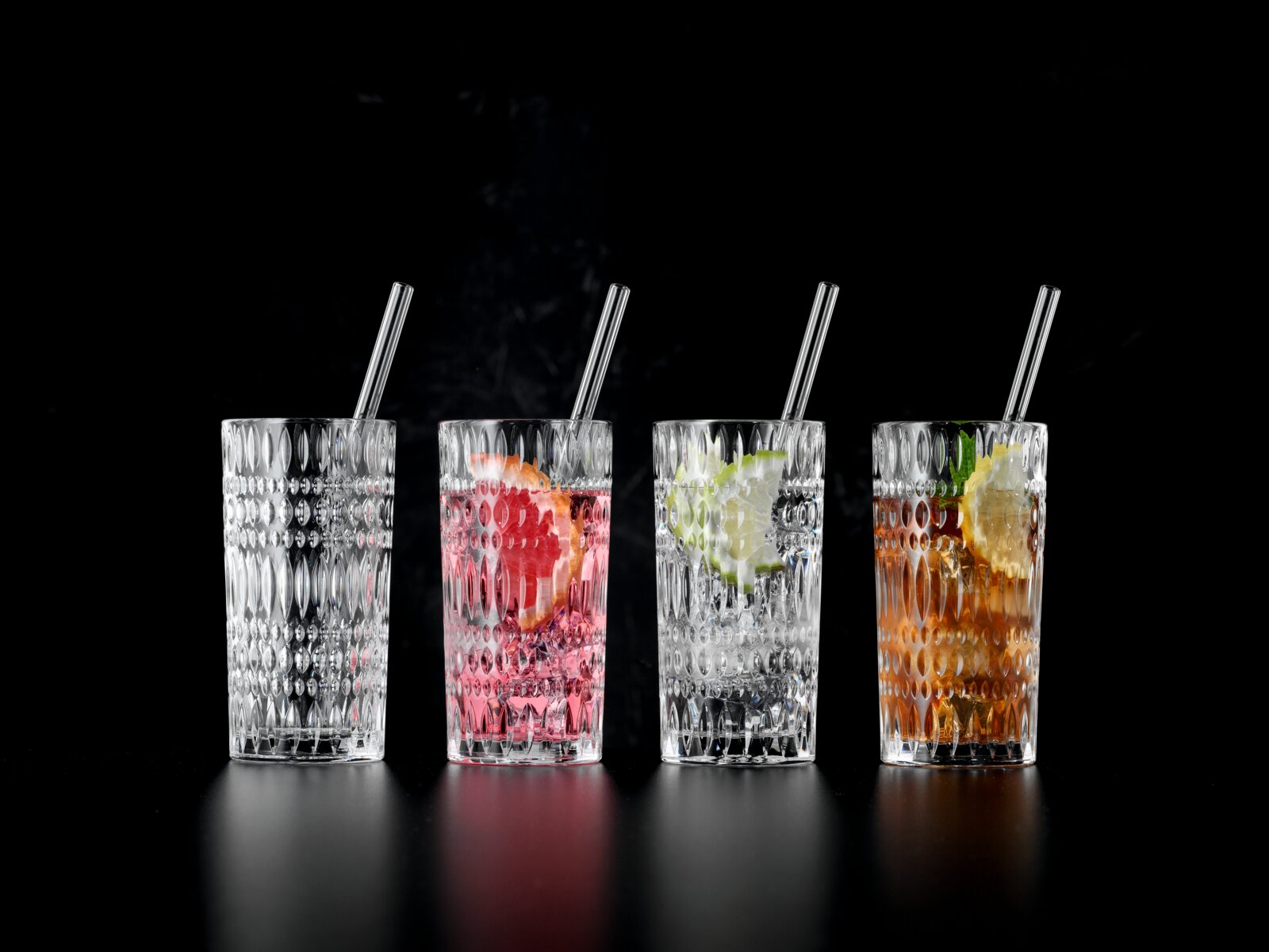 Vier hoge, facetgeslepen longdrinkglazen met rietjes op zwarte achtergrond één leeg en drie met cocktails, ijs en citrus, gefotografeerd in frontaal aanzicht op ooghoogte.