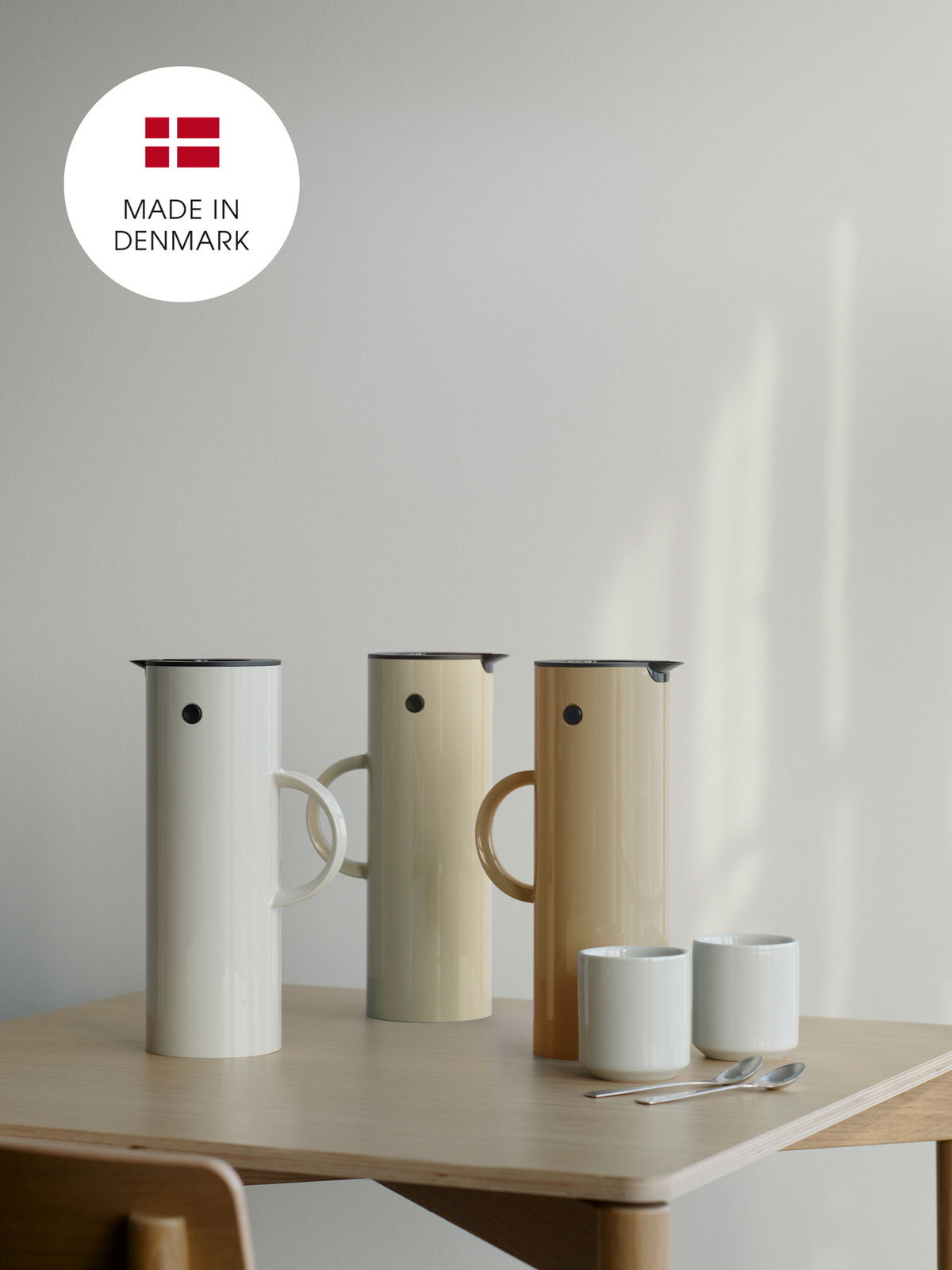 Stelton Isolierkanne EM77 Drei Isolierkannen in verschiedenen Farben auf einem Tisch, mit der Aufschrift 'Made in Denmark' im Hintergrund. Perspektive von vorne.