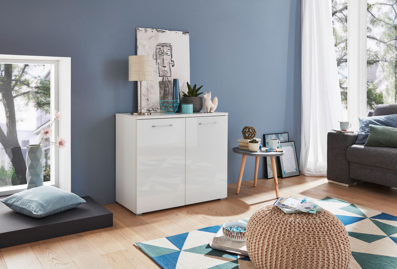 Weißes Sideboard mit zwei Türen, dekoriert mit Pflanzen und Kunstobjekten, in einem modernen Wohnzimmer mit blauer Wand und Holzboden. Perspektive von vorne.