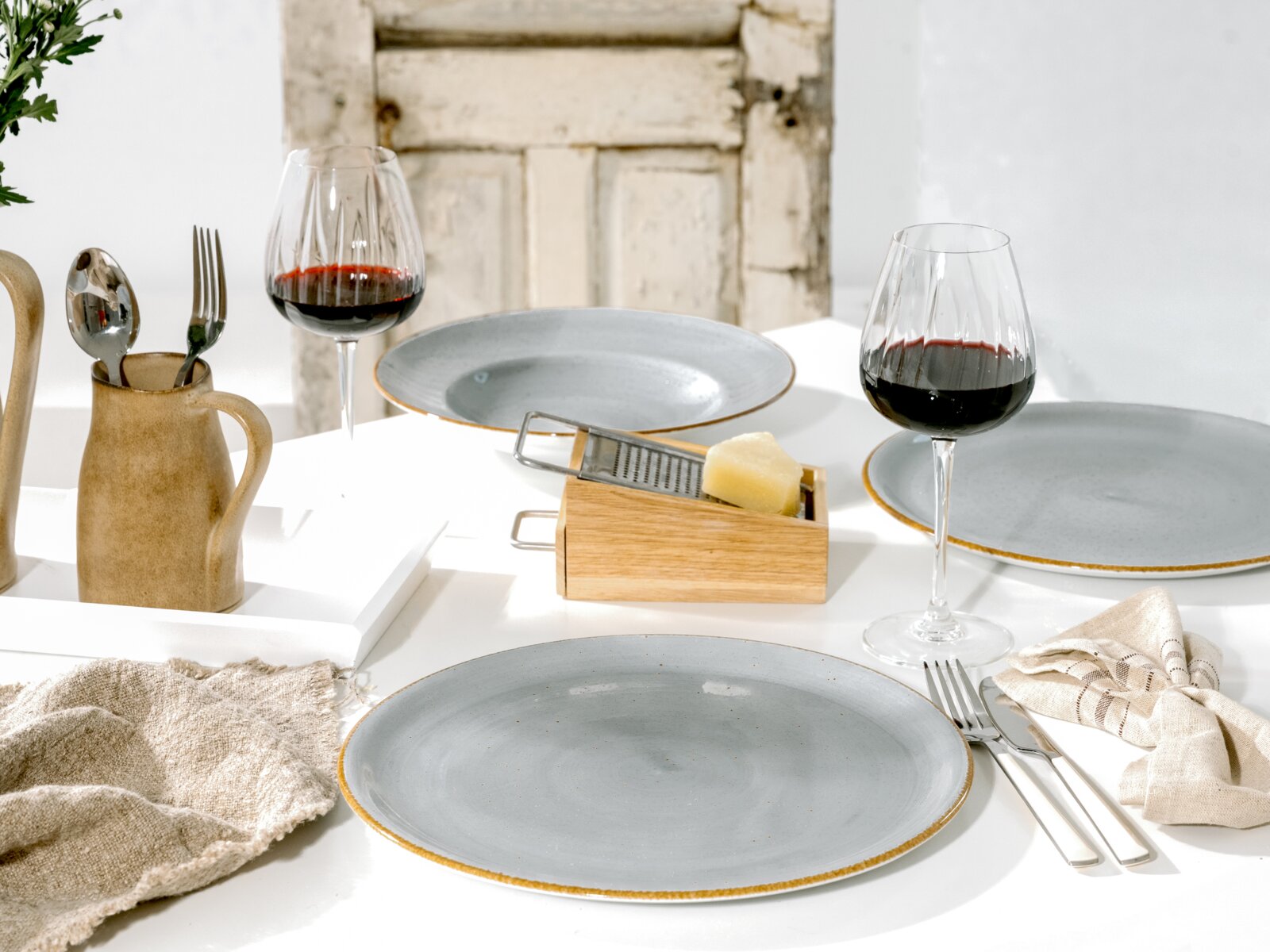 Gedeckter Esstisch mit grauen Keramiktellern, zwei Rotweingläsern, Besteck, Leinenserviette und Holzreibe mit Käse, aufgenommen aus leicht erhöhter schräger Perspektive.