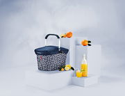 Tragetasche mit dunkelblauer Abdeckung und geometrischem Muster, umgeben von Orangen und Zitronen, auf weißen Podesten aus seitlicher Perspektive.