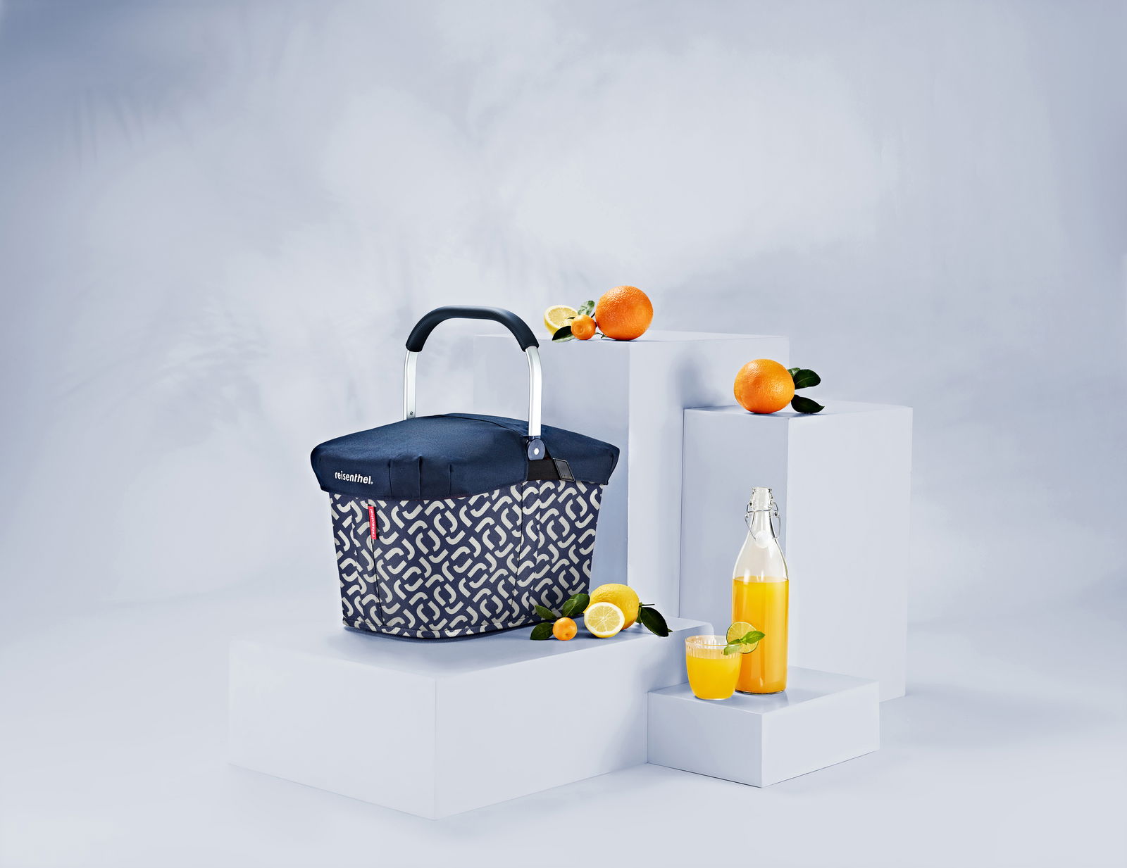 Tragetasche mit dunkelblauer Abdeckung und geometrischem Muster, umgeben von Orangen und Zitronen, auf weißen Podesten aus seitlicher Perspektive.