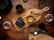 Draufsicht auf ein rustikales Holz-Servierbrett mit schwarzer Kaffeetasse und Espresso, daneben goldfarbene Löffel, weitere schwarze Becher und Wassergläser auf dunklem Holztisch.