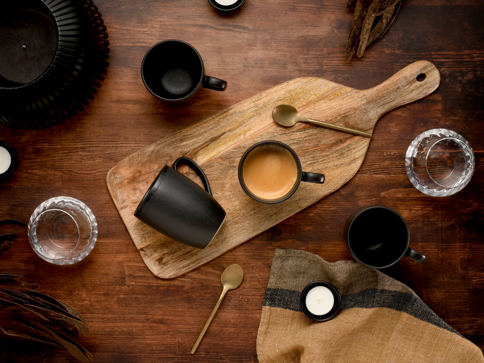 Draufsicht auf ein rustikales Holz-Servierbrett mit schwarzer Kaffeetasse und Espresso, daneben goldfarbene Löffel, weitere schwarze Becher und Wassergläser auf dunklem Holztisch.