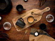 Draufsicht auf ein rustikales Holz-Servierbrett mit schwarzer Kaffeetasse und Espresso, daneben goldfarbene Löffel, weitere schwarze Becher und Wassergläser auf dunklem Holztisch.