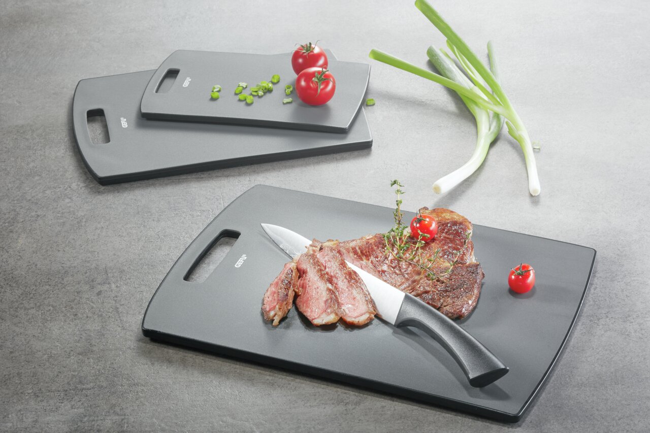 Drei graue Schneidbretter in verschiedenen Größen, auf einem liegt ein Messer mit geschnittenem Fleisch und Kirschtomaten, fotografiert aus der Vogelperspektive.