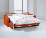 Orangefarbenes Schlafsofa mit ausgeklapptem Metallgestell, Matratze und weißer Bettwäsche mit rotem Schriftmuster, aus schräger Vorderansicht