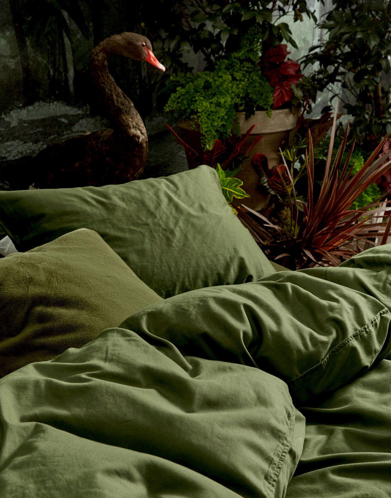 Grüne Bettwäsche in einem natürlichen Ambiente mit Pflanzen und einem schwarzen Schwan im Hintergrund, seitliche Perspektive.