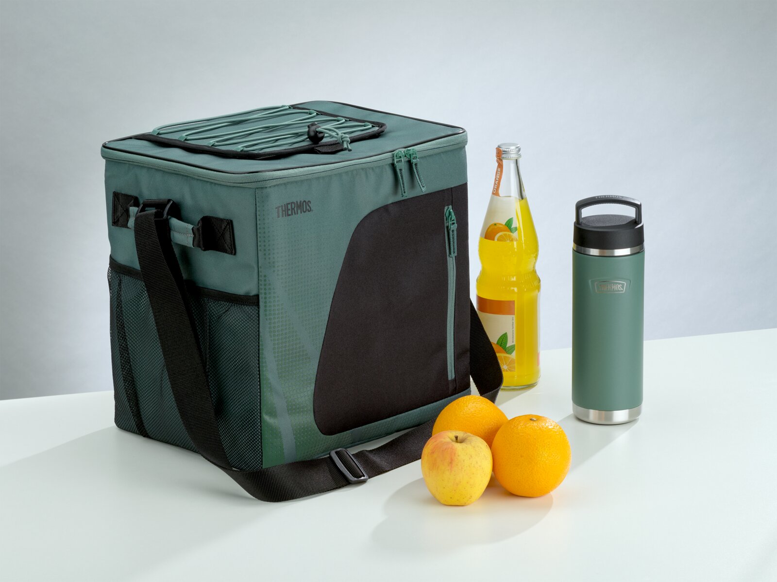 Grün-schwarze THERMOS Kühltasche mit Schultergurt und elastischer Deckelkordel, in schräger Vorderansicht neben Thermobecher, Getränkeflasche und frischem Obst präsentiert