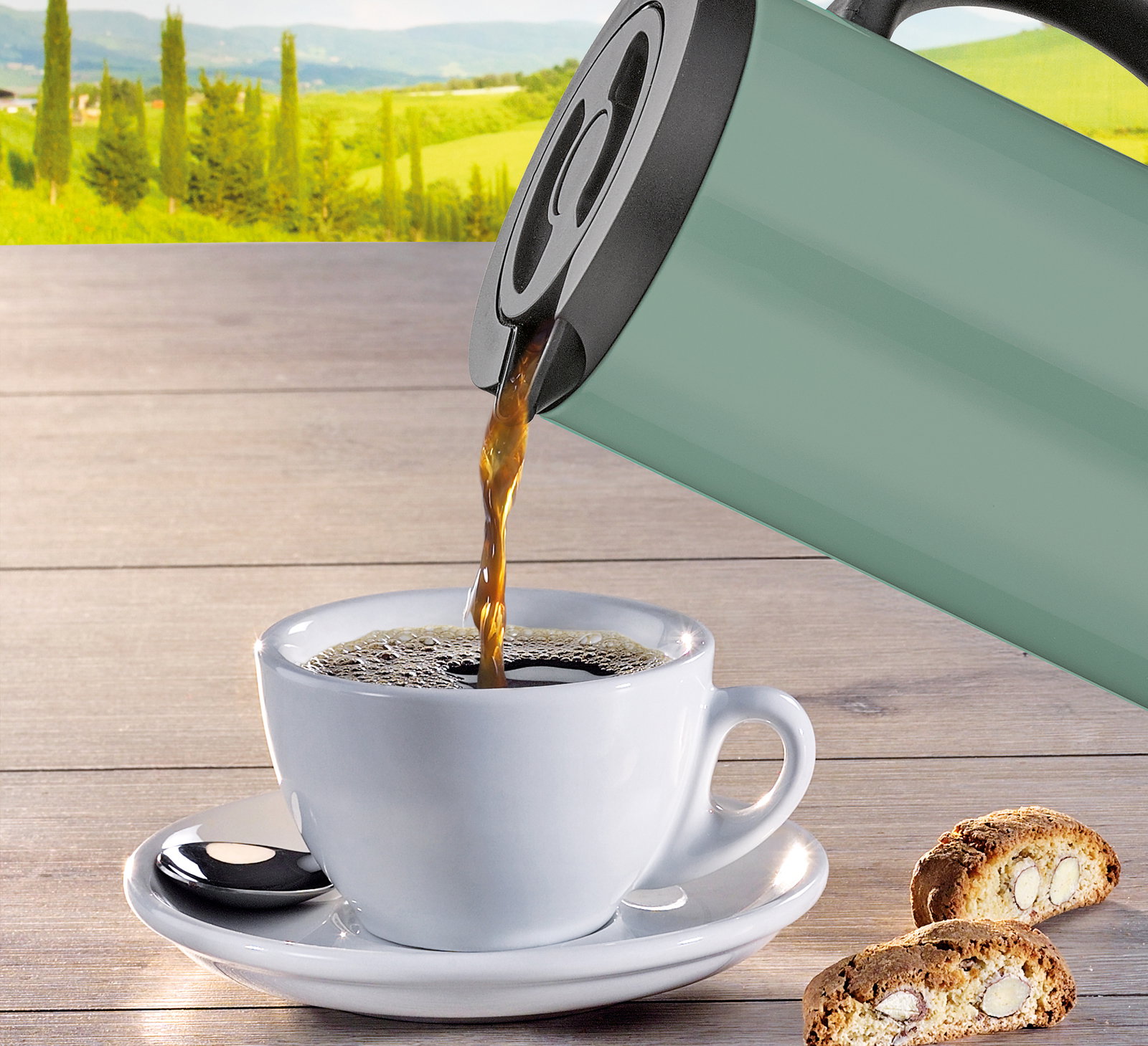 Isolierkanne in Grün, die Kaffee in eine weiße Tasse auf einem Holztisch gießt, mit Keksen im Vordergrund und einer Landschaft im Hintergrund, seitliche Perspektive.