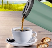Isolierkanne in Grün, die Kaffee in eine weiße Tasse auf einem Holztisch gießt, mit Keksen im Vordergrund und einer Landschaft im Hintergrund, seitliche Perspektive.