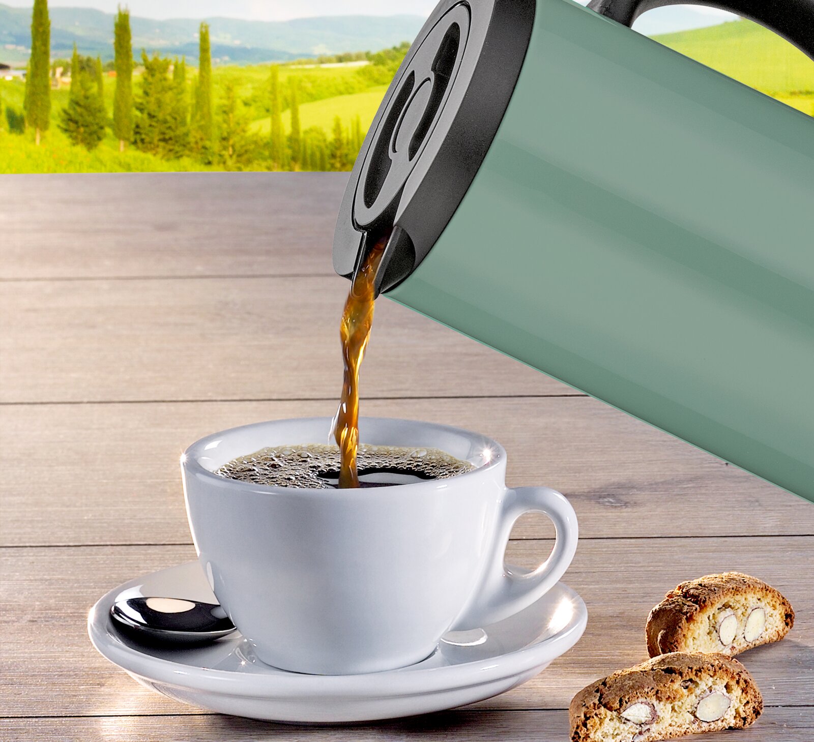 Isolierkanne in Grün, die Kaffee in eine weiße Tasse auf einem Holztisch gießt, mit Keksen im Vordergrund und einer Landschaft im Hintergrund, seitliche Perspektive.