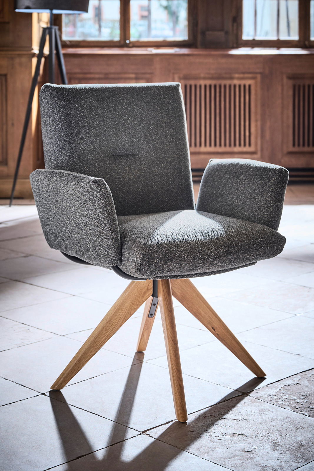 Grauer Armlehnstuhl mit Holzbeinen, fotografiert aus einem leicht seitlichen Winkel in einem Raum mit Holzwänden und großen Fenstern.