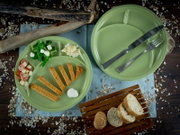 Zwei grüne, runde Menüteller mit Trennfächern, einer mit Besteck und einer mit servierten Speisen, auf rustikalem Holztisch mit Brot und Dekostreu, fotografiert aus der Vogelperspektive (Draufsicht).