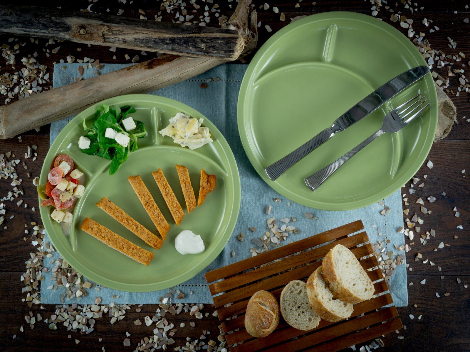 Zwei grüne, runde Menüteller mit Trennfächern, einer mit Besteck und einer mit servierten Speisen, auf rustikalem Holztisch mit Brot und Dekostreu, fotografiert aus der Vogelperspektive (Draufsicht).
