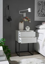 Moderne Nachtkommode mit zwei Schubladen, weiß mit geriffelter Front, auf schwarzem Metallgestell. Perspektive von vorne rechts.