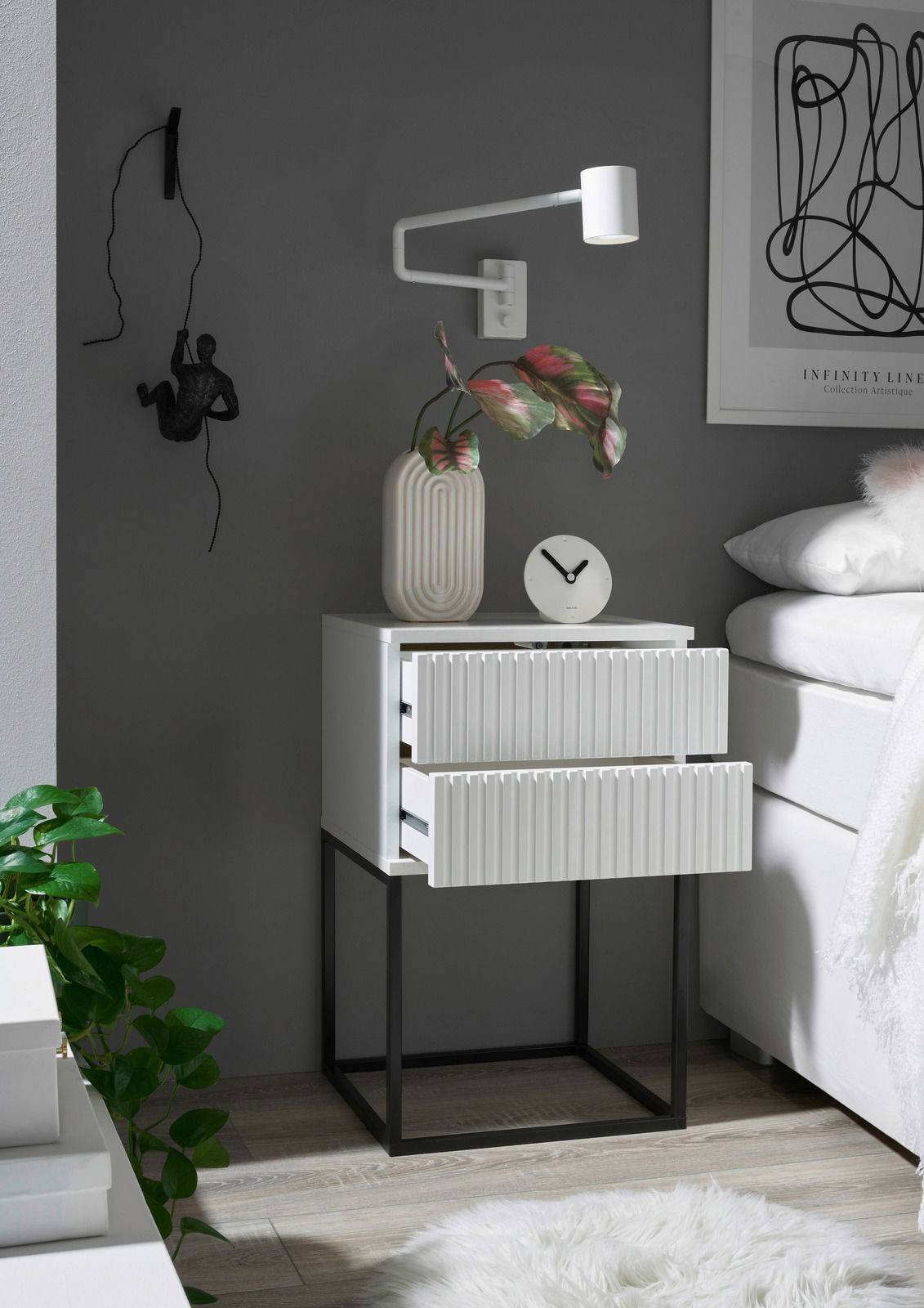 Moderne Nachtkommode mit zwei Schubladen, weiß mit geriffelter Front, auf schwarzem Metallgestell. Perspektive von vorne rechts.