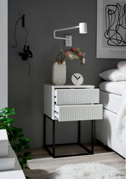 Moderne Nachtkommode mit zwei Schubladen, weiß mit geriffelter Front, auf schwarzem Metallgestell. Perspektive von vorne rechts.